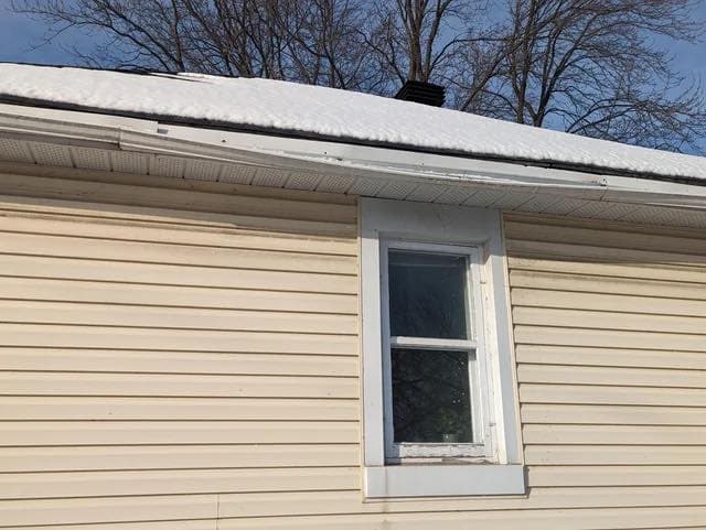 Snow-covered roofline of a beige house with a single window and bare trees in the background.