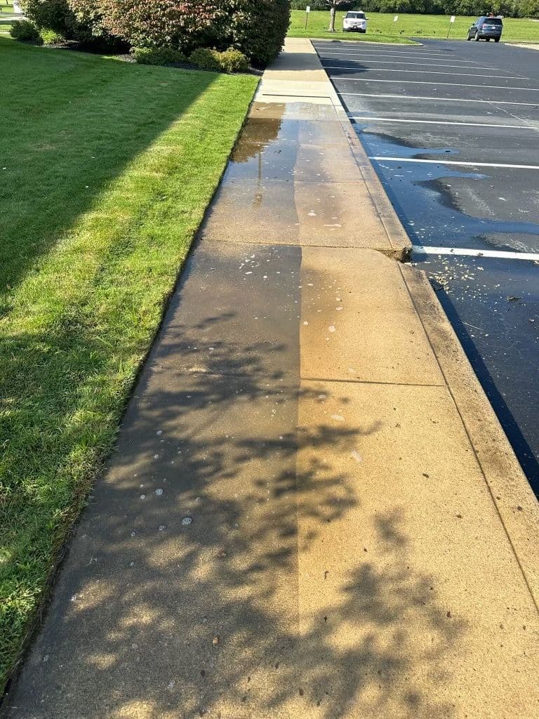 Water pooling on sidewalk beside grassy area and empty parking lot under clear sky.