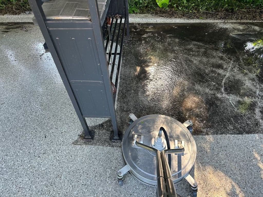 Pressure washer cleaning pavement near a black metal grill with water splashes.