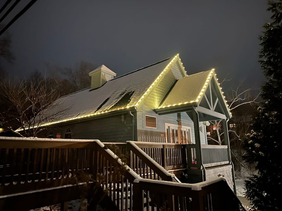 Snow-covered house at night adorned with warm string lights, surrounded by trees.